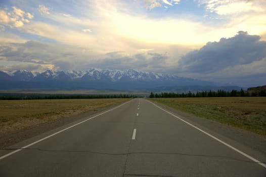 A direct asphalt road abutting against mountain ranges covered with snow under a cloudy sky. North Chuy Range. Altai, Siberia, Russia.