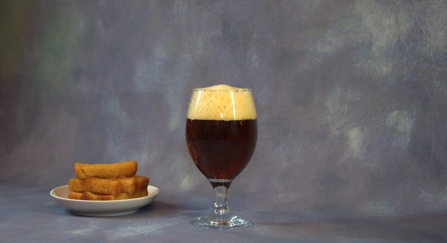 A full glass of dark beer with foam and a plate with wheat garlic croutons on a gray background. Close-up.