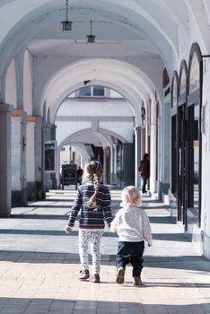 Two young children walk together hand in hand in the old town arcade street. Family love and friendship theme.