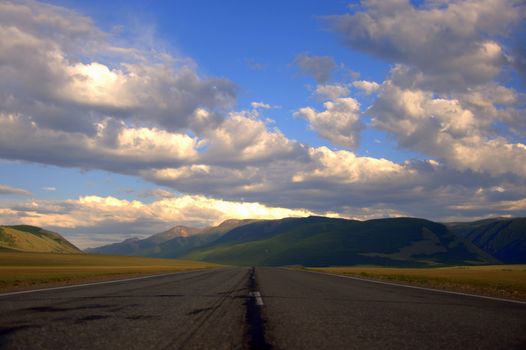 Cracked asphalt road running through the valley to the snowy ridges. Kurai steppe, Altai, Siberia, Russia.