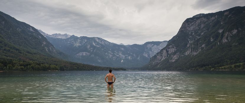 Man going to swim in freezing cold lake Bohinj, Alps mountains, Slovenia, on tranquil overcast morning.