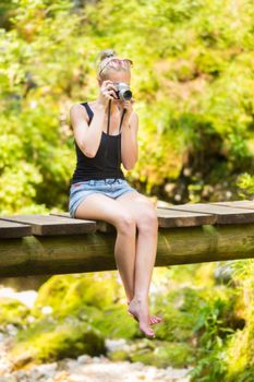 Beautiful barefooted blonde caucasian girl wearing jeans shorts an sporty black sleeveless t-shirt, sitting on a vintage wooden bridge, taking photos with retro camera.