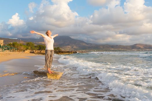 Relaxed woman, arms rised, enjoying sun, freedom and life an beautiful beach in sunset. Young lady feeling free, relaxed and happy. Concept of vacations, freedom, happiness, enjoyment and well being.