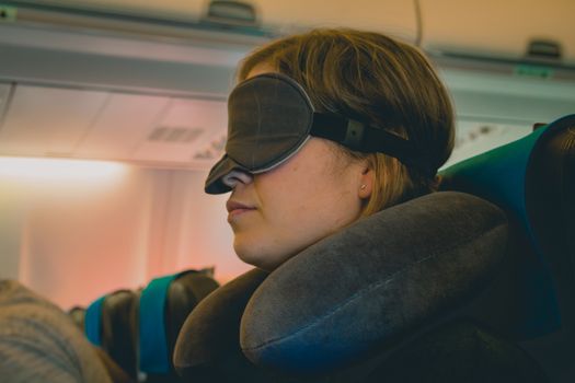 Tired caucasian blond woman with neck pillow and blindfold sleeping on a seat while traveling in a commercial airplane.