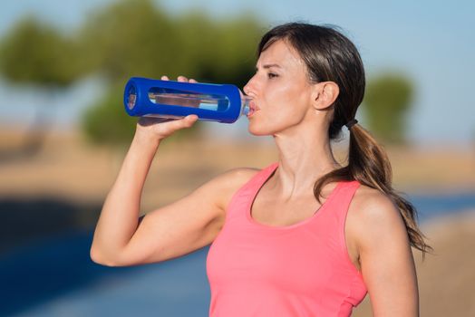 Side view of beautiful girl in sport clothes drinking water after workout on the park