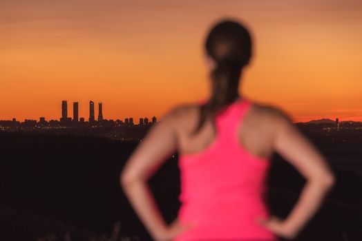 Woman on sport clothes watching sunset over city skyline. Focus is on background.