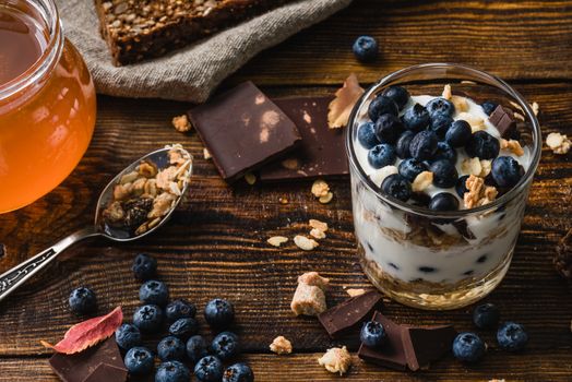 Granola with yogurt, blueberries, honey and chocolate bar on wooden table with ingredients.