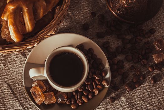 A cup of coffee on textile with coffee beans, dark candy sugar, pots, basket and cake