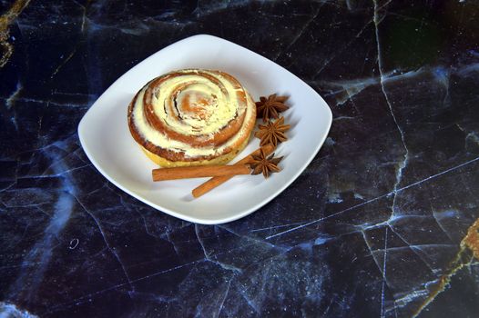 White ceramic plate with a bun in the glaze, anise and cinnamon on a marble blue table. Close-up.