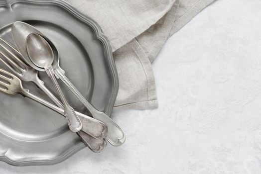 Various silverware on a pewter plate and gray flax napkin are on the background of gray concrete surface, with copy-space