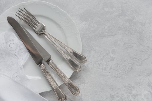 Silver cutlery on a porcelain plate and white napkin with Belgian lace are on the background of gray concrete surface, with copy-space