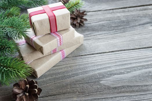 Christmas composition of pine cones, spruce branches and stack of gift boxes tied with red ribbons on the background of old unpainted wooden boards; with copy-space