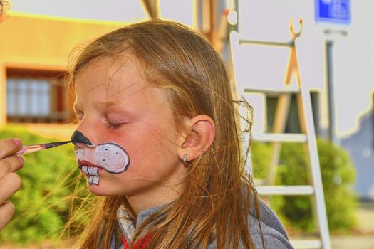 Woman painting face of kid outdoors. Baby face painting. Little girl getting her face painted like a rabbit by face painting artist.