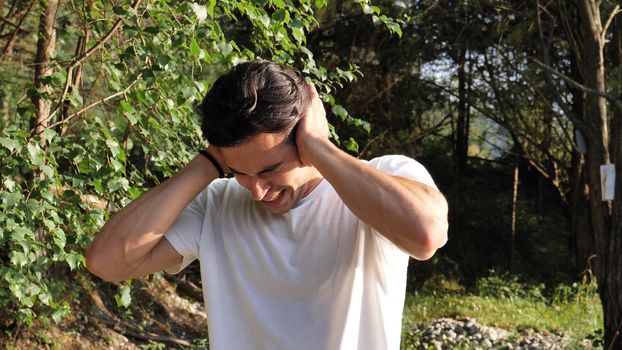 Young handsome man standing in nature, covering ears in pain and frustration because of too much loud noise