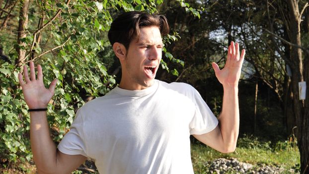 Young handsome man standing in nature, covering ears in pain and frustration because of too much loud noise