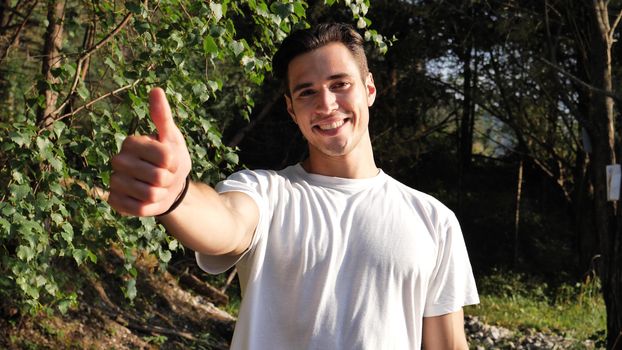 Young handsome man standing in nature, happy, doing thumb up sign with one hand