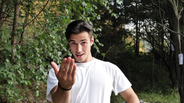 Young handsome man standing in nature, saying Come Here or Come Closer with hand gesturing