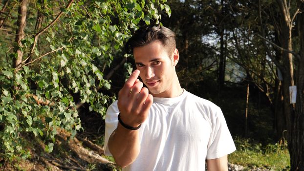 Young handsome man standing in nature, saying Come Here or Come Closer with hand gesturing