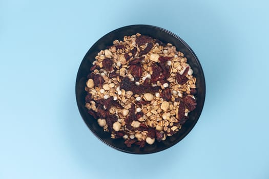 A fresh granola with dried and candied nuts and fruits in black bowl on blue bakground. Concept of nutrient and healty breakfast or meal