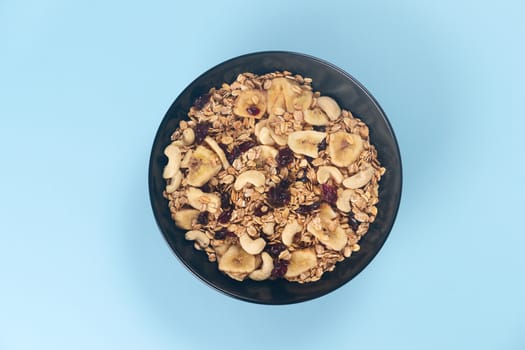 A fresh granola with dried and candied nuts and fruits in black bowl on blue bakground. Concept of nutrient and healty breakfast or meal