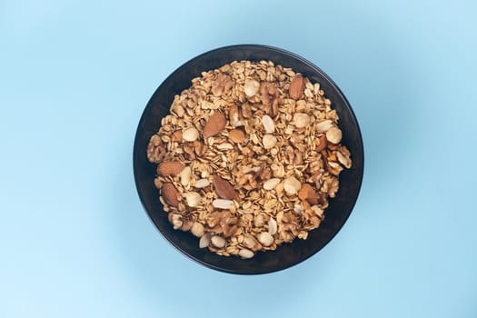 A fresh granola with dried and candied nuts and fruits in black bowl on blue bakground. Concept of nutrient and healty breakfast or meal