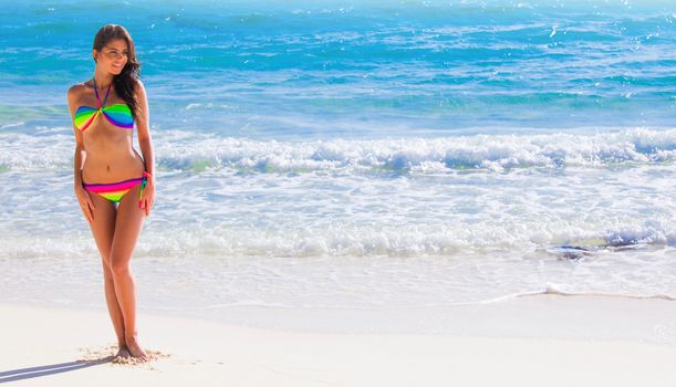 Happy tanned girl in rainbow bikini at seaside, blue sea water in background