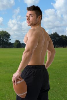 Muscular american football player standing with ball in hand, seen from the back, in playing field