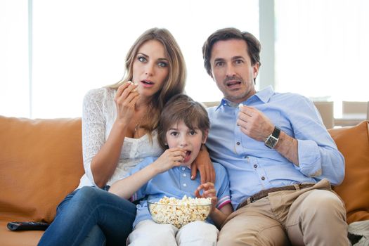 Beautiful young parents and their son are watching TV, eating popcorn sitting on couch at home