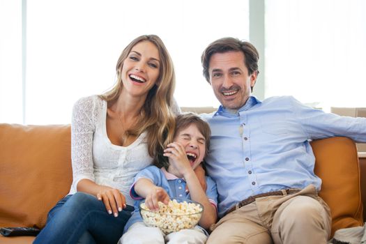 Beautiful young parents and their son are watching TV, eating popcorn and smiling while sitting on couch at home