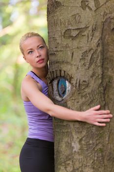 Relaxed young lady embracing a tree receiving life energy from the nature. Eye carved in tree trunk.