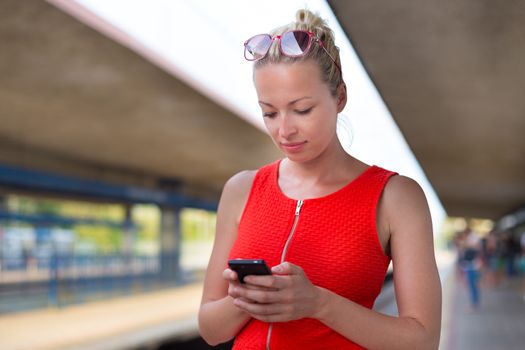 Young woman in red dress with a cell phone in her hand waiting on the platform of a railway station chacking train schedule online.