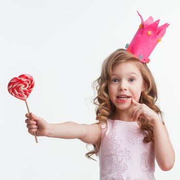 Beautiful little candy princess girl in crown holding big pink heart lollipop and smiling isolated on white