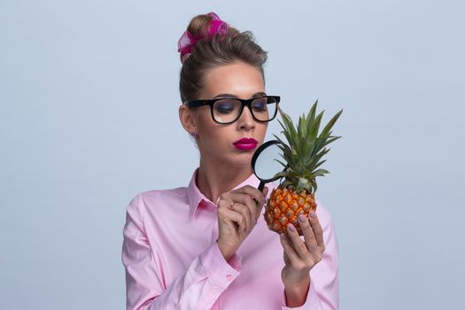 Blonde woman in glasses holding pineapple and look at it through magnifying glass investigating quality and usefulness