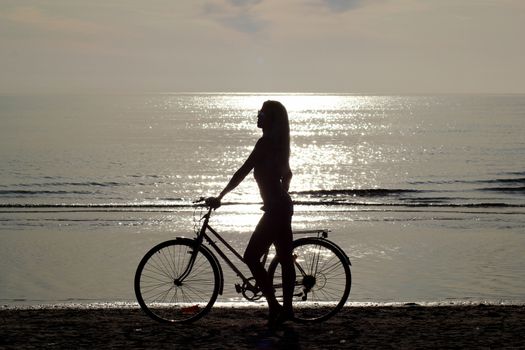 silhouette of a girl with long hair on a bicycle riding on the sand by the sea. Against the background of a small surf of the Baltic Sea. Shooting against the sun. Reflection of the sun on the surface of the water.