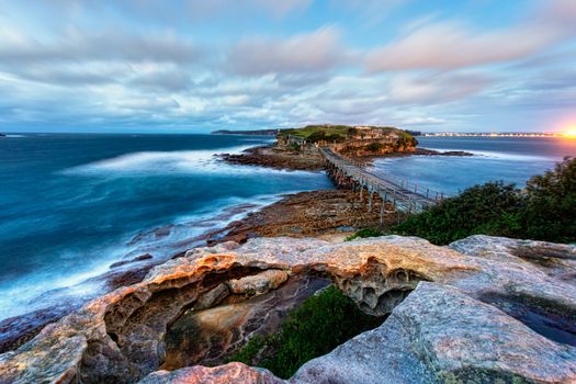 Strong winds whip up the ocean and move the clouds briskly across the sky.  A long exposure exaggerates this movement.  Views across the old timber bridge to Bare Island in the dawn hours 