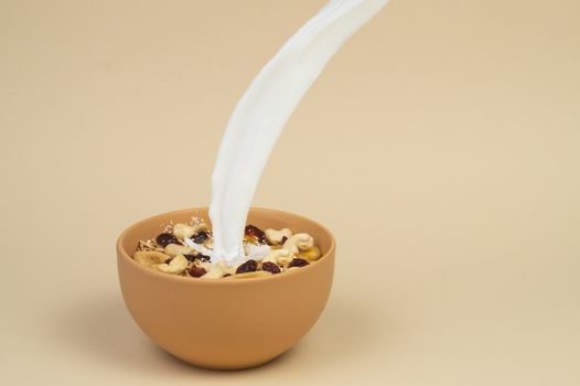 A stream og milk pours onto granola with dried and candied fruits and nuts in beige colour bowl. Bowl on a beige background. Concept of nutrient and healty breakfast or meal