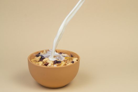 A stream og milk pours onto granola with dried and candied fruits and nuts in beige colour bowl. Bowl on a beige background. Concept of nutrient and healty breakfast or meal