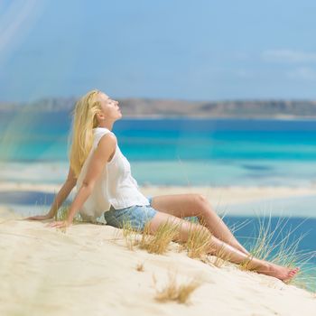 Relaxed woman enjoying sun, freedom and life an a beautiful sandy beach of Balos in Greece. Young lady feeling free, relaxed and happy. Vacations, freedom, happiness, enjoyment and well being.