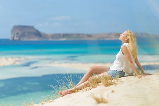 Relaxed woman enjoying sun, freedom and life an a beautiful sandy beach of Balos in Greece. Young lady feeling free, relaxed and happy. Vacations, freedom, happiness, enjoyment and well being.