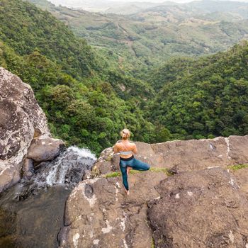 Active sporty woman relaxing in nature, practicing yoga on high clif by 500 feet waterfall at Black river gorges national park on tropical paradise island of Mauritius.