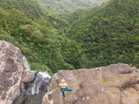 Active sporty woman relaxing in nature, practicing yoga on high clif by 500 feet waterfall at Black river gorges national park on tropical paradise island of Mauritius.