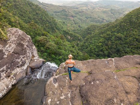 Active sporty woman relaxing in nature, practicing yoga on high clif by 500 feet waterfall at Black river gorges national park on tropical paradise island of Mauritius.