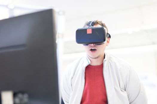 Young man wearing virtual reality headset and gesturing while sitting at his desk in creative office. Virtual reality in video games.