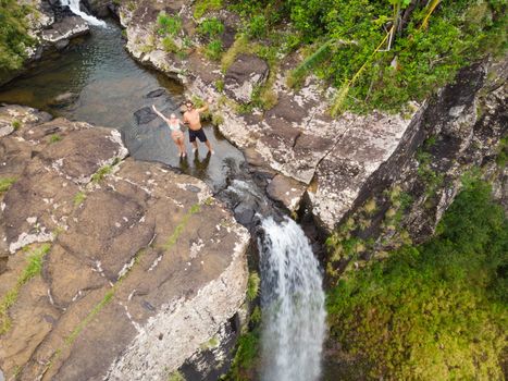 Aerial top view of travel couple waving to drone, standing on the edge of 500 feet waterfall in the tropical island jungle of Black river gorges national park on Mauritius island.