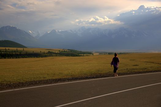 A young girl in a tight-fitting dress walks along the edge of the road against the backdrop of a valley at the foot of snowy mountain ranges under dramatic thunderclouds. Altai, Siberia, Russia.