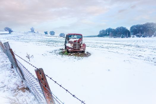 A rusty old vintage car sits in a snow covered field in winter.   There is a leaning rusty barbed wire fence in the foreground .   Space for copy