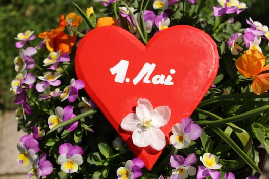 The picture shows a red wooden heart in flowers with the text may day.