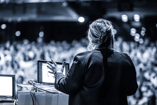 Female speaker giving a talk on corporate business conference. Unrecognizable people in audience at conference hall. Business and Entrepreneurship event. Black and white, blue toned image.