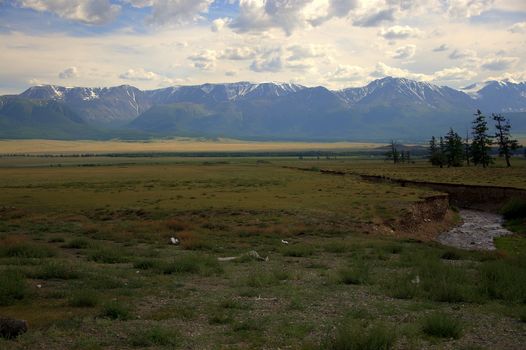 A small river flowing in a canyon across the steppe to meet the snow-capped mountain peaks. Altai, Siberia, Russia.