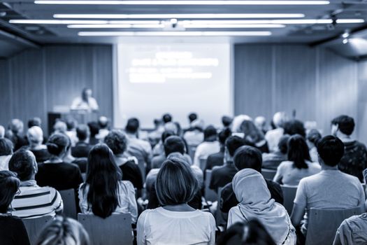 Speaker giving a talk in conference hall at business event. Audience at the conference hall. Business and Entrepreneurship concept. Focus on unrecognizable people in audience. Blue toned.